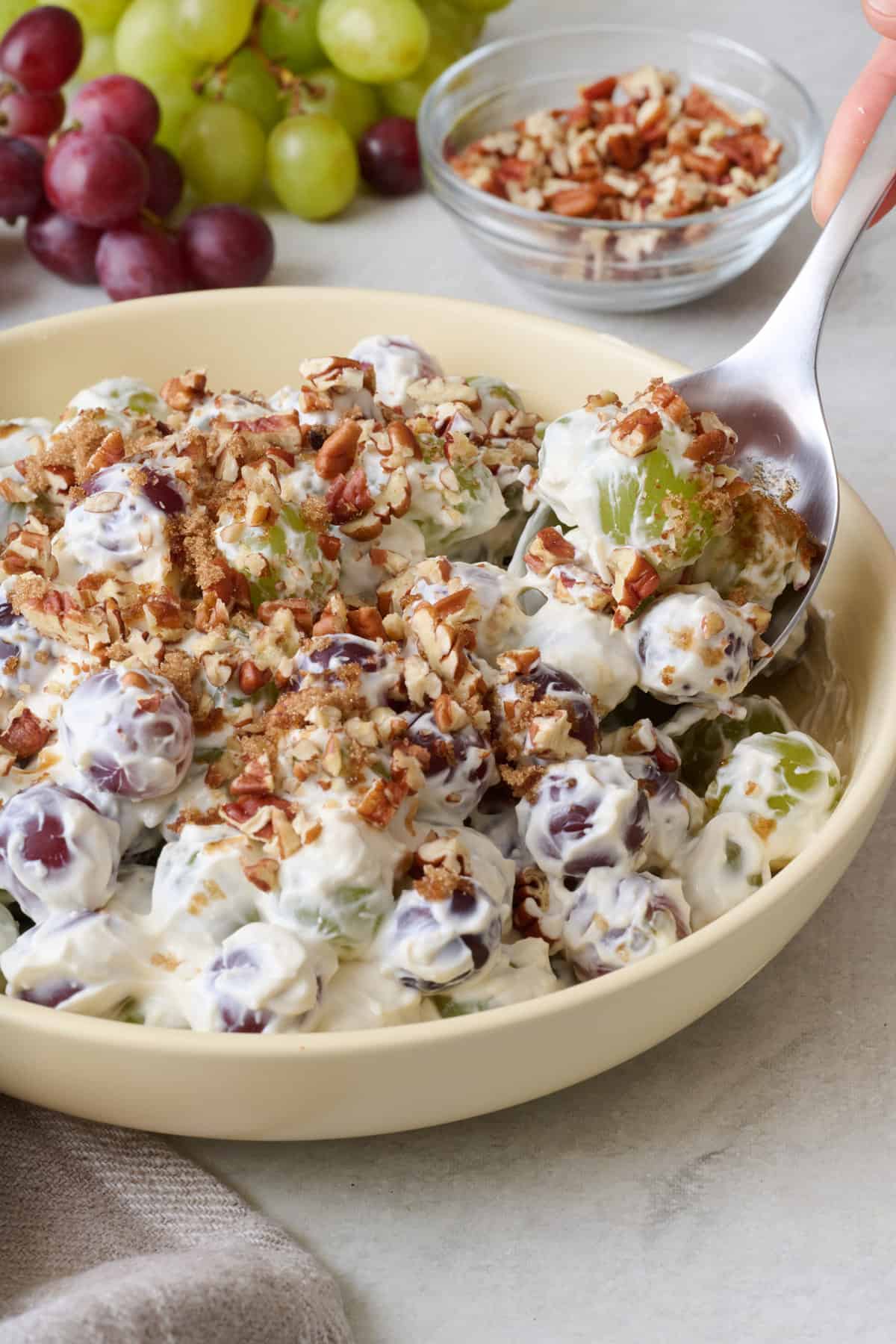 Spoon scooping grape salad with chopped pecans from a bowl with fresh grapes and bowl of chopped pecans nearby.