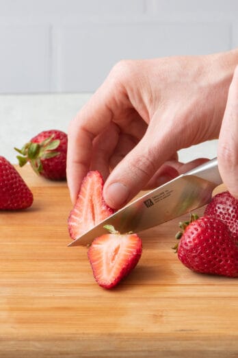 How to cut strawberries: Hand slicing strawberry in half with a paring knife on wooden cutting board.