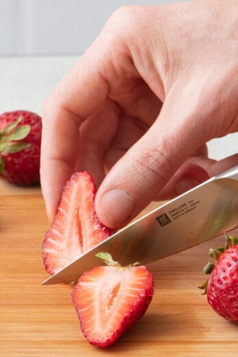 Hand slicing strawberry in half with a paring knife on wooden cutting board.
