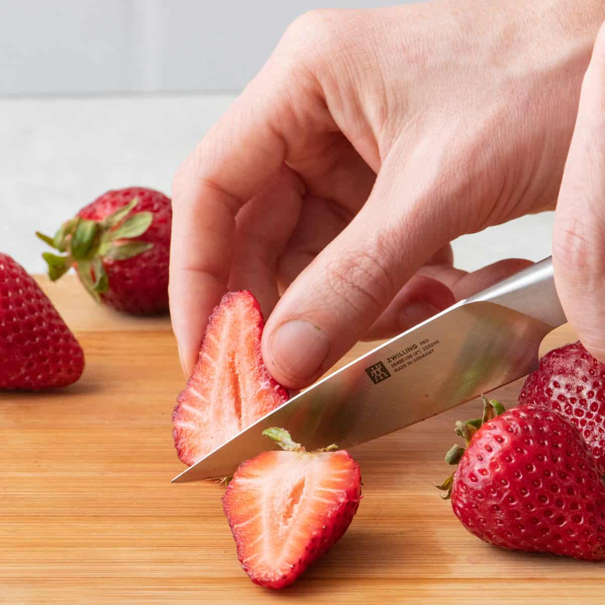 Hand slicing strawberry in half with a paring knife on wooden cutting board.