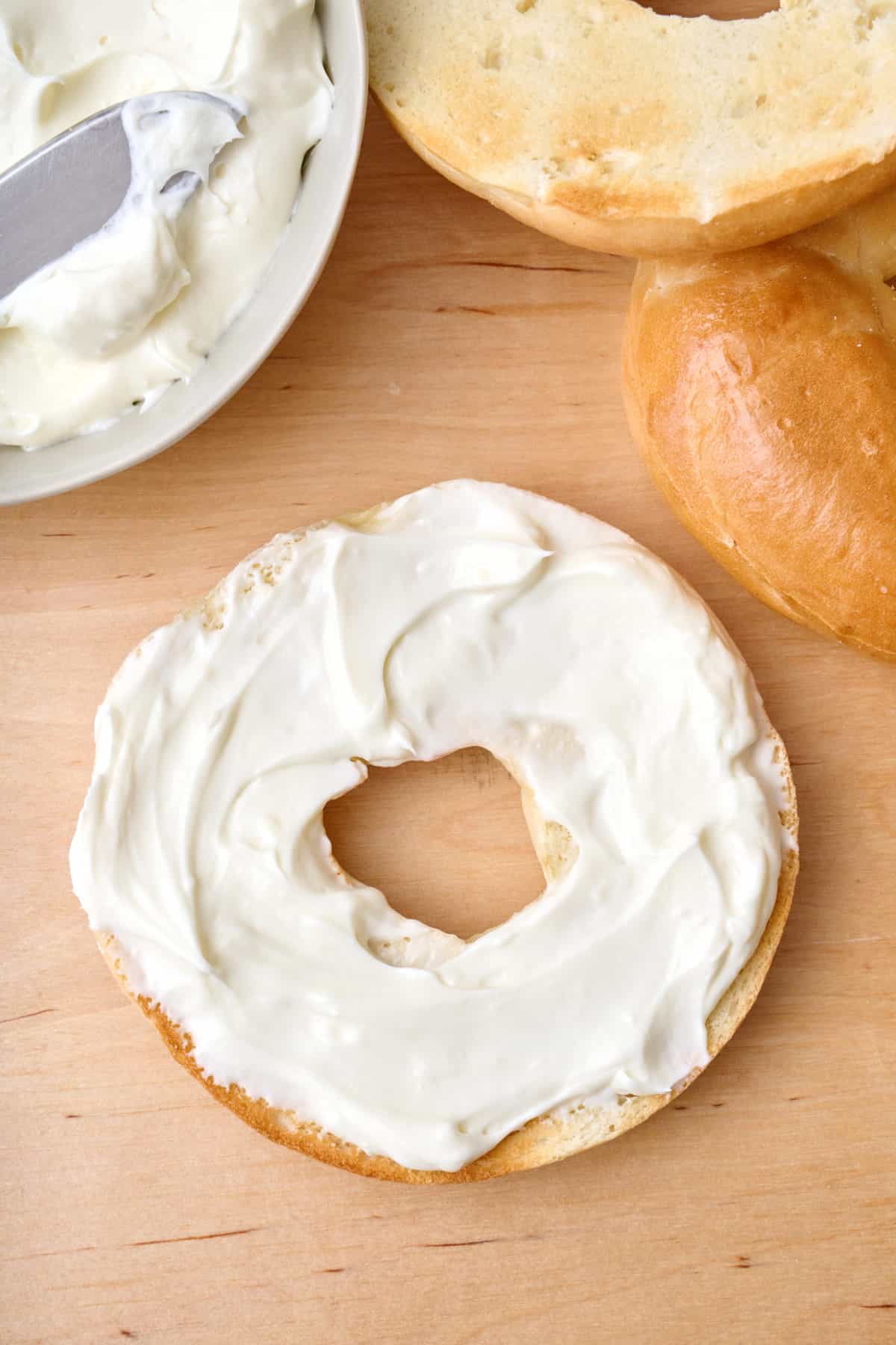 Whipped cream cheese on a bagel with more cream cheese and toasted bagels nearby.