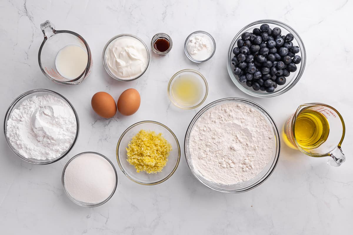 Two eggs, measuring cups of oil and milk, and bowls containing blueberries, flour, baking powder and salt, Greek yogurt, vanilla, lemon zest, sugar, lemon juice, and powdered sugar on a marble countertop.