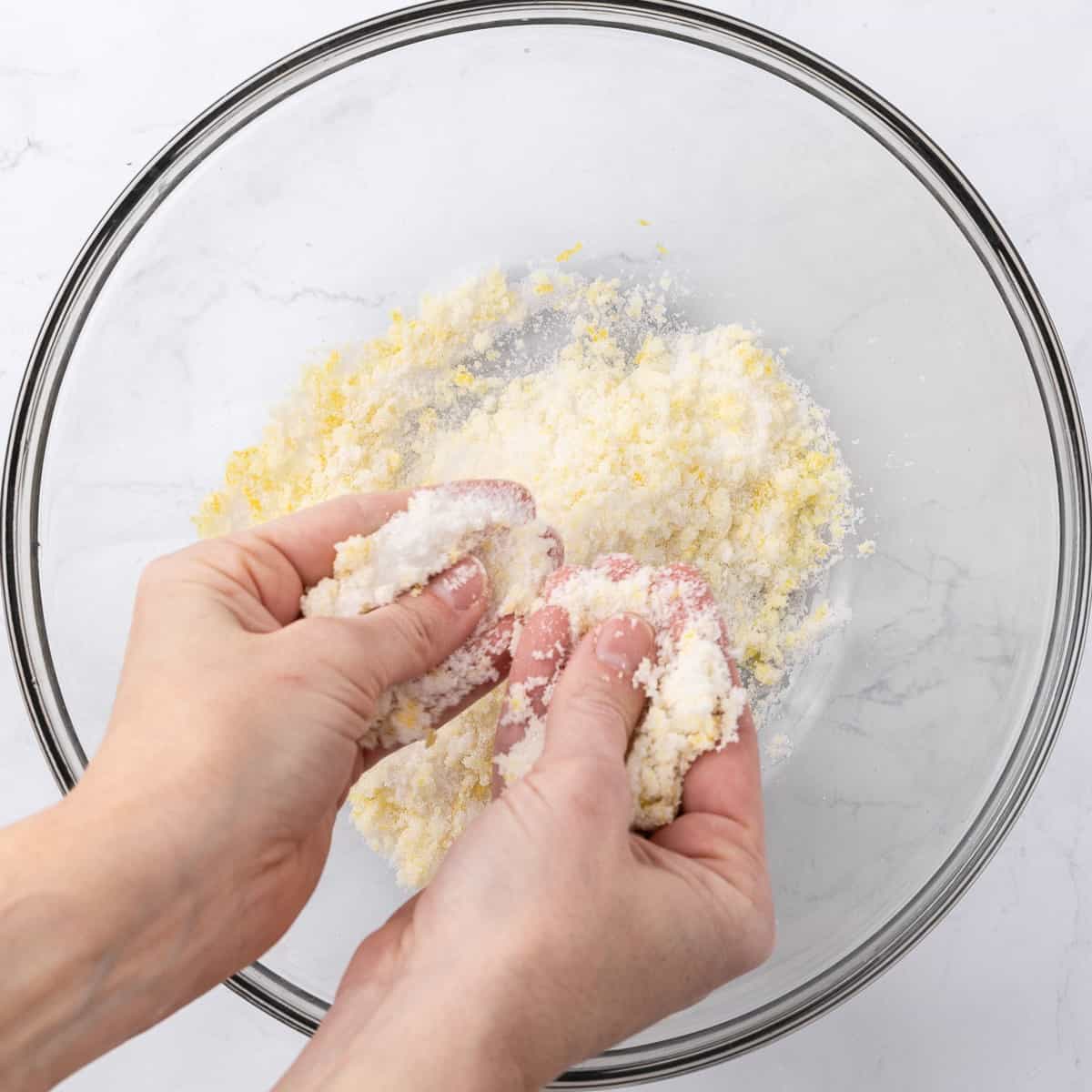 A person using their hands to rub granulated sugar and lemon zest together in a large glass mixing bowl.