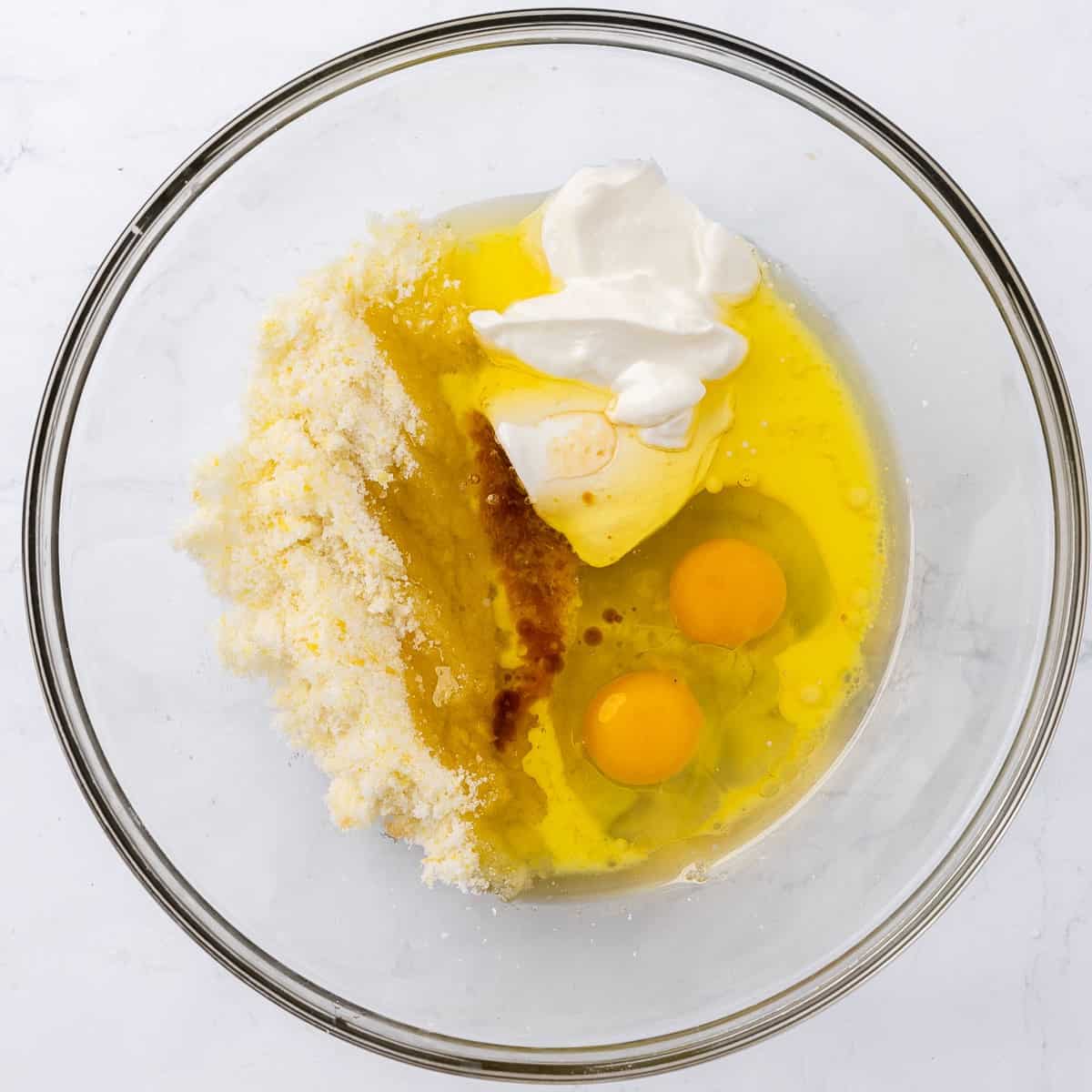 Sugar, lemon zest, eggs, Greek yogurt, olive oil, milk, vanilla, and lemon juice in a large glass mixing bowl.