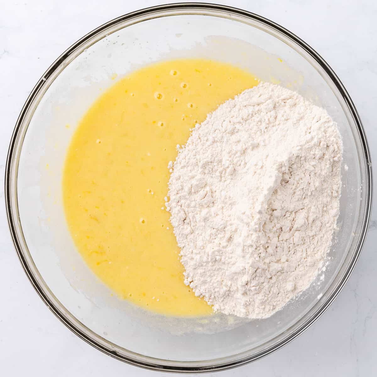Flour mixed with baking powder and salt on top of wet ingredients in a large glass mixing bowl.