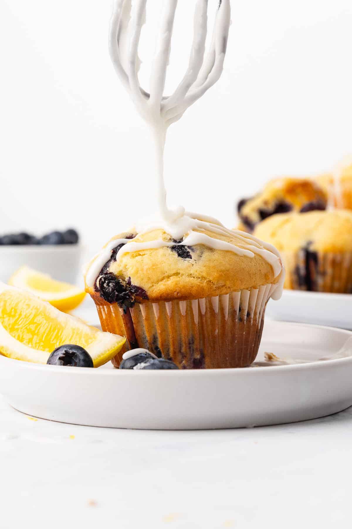 A whisk drizzling glaze onto a lemon blueberry muffin on a plate.