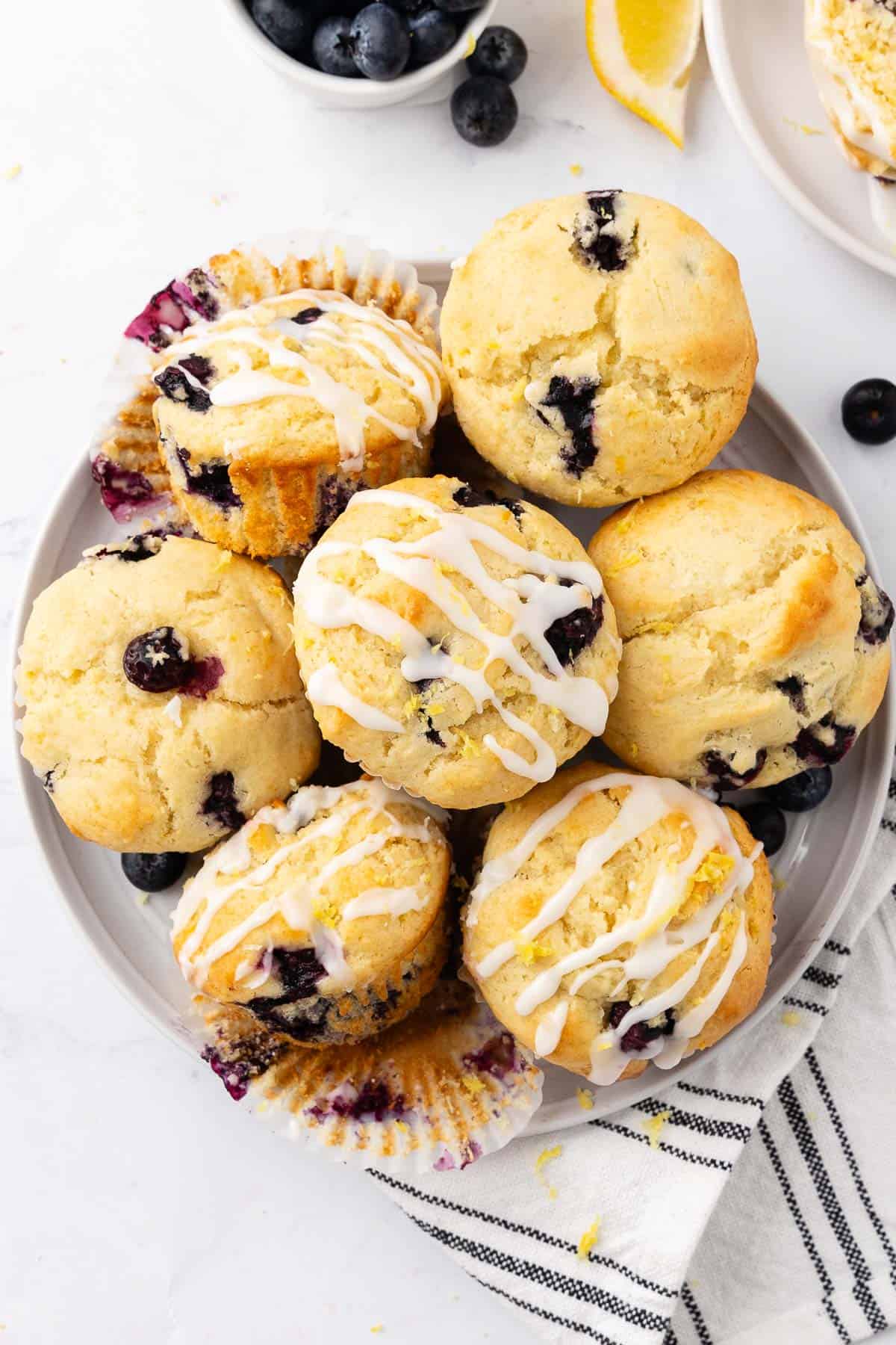 Several Lemon Blueberry Muffins on a plate. Some of the muffins are topped with a lemon glaze.