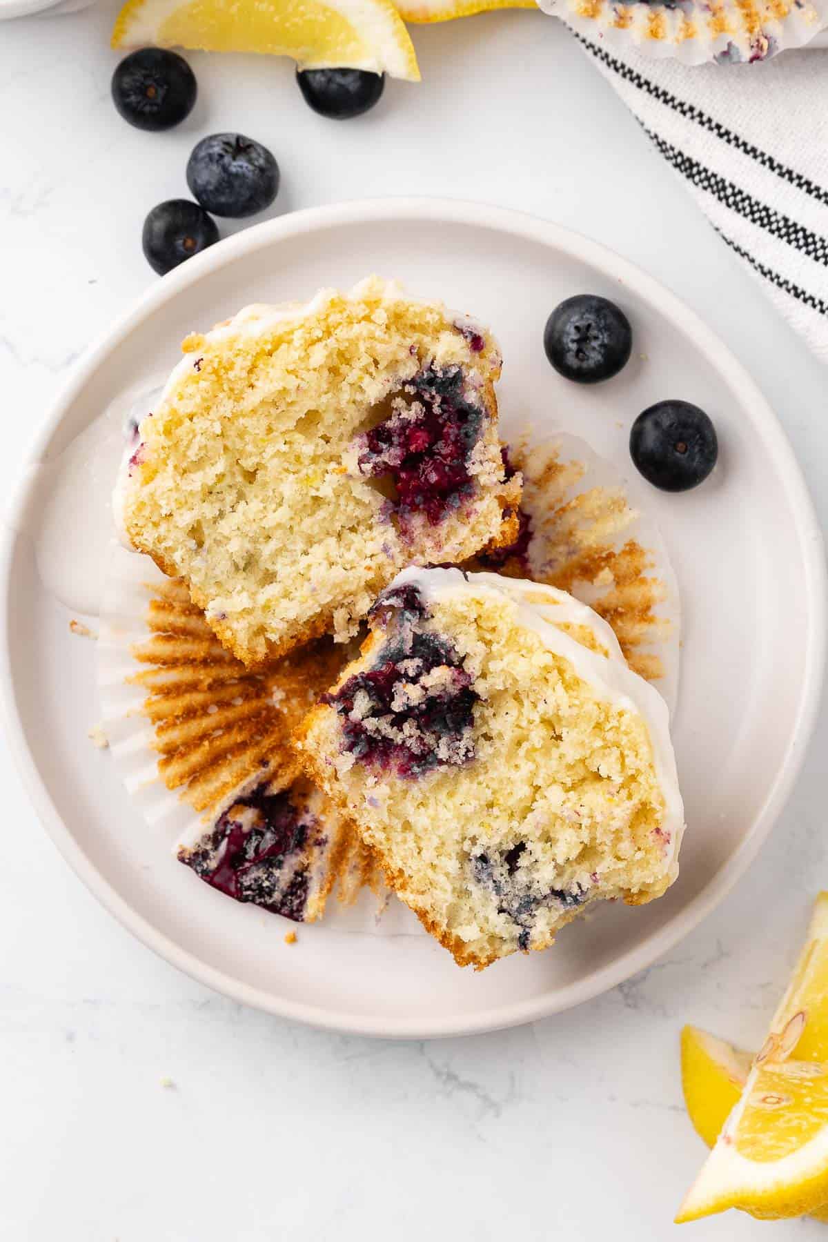 A lemon blueberry muffin topped with a lemon glaze that's split in half on a small white plate.