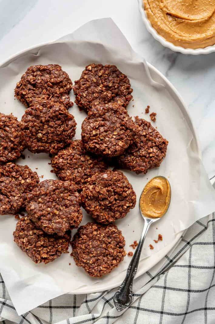 Parchment paper-lined plate with batch of no-bake oatmeal cookies and spoonful of peanut butter with more peanut butter and linen napkin nearby.