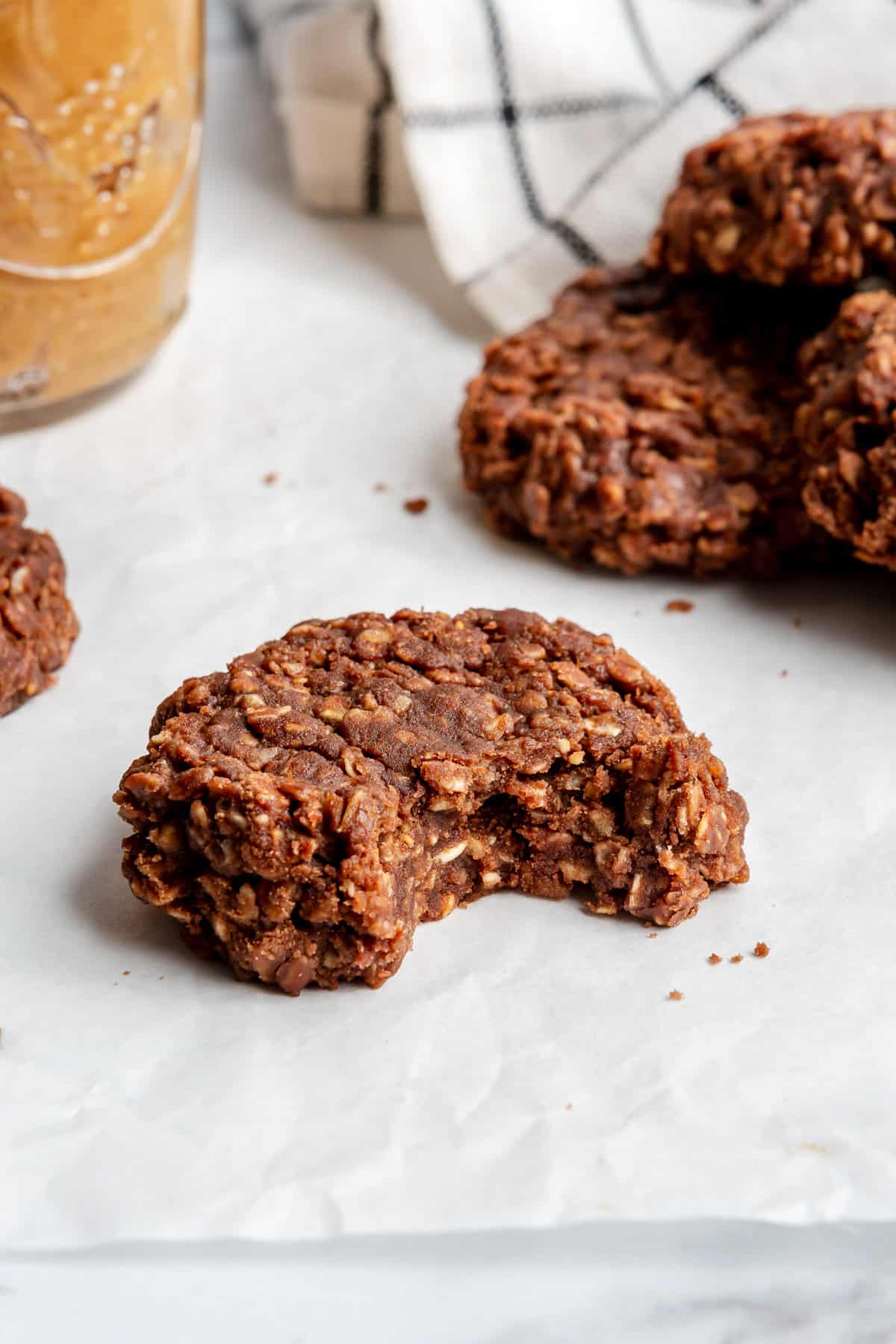 No-bake oatmeal cookie with a bite taken out on parchment paper with other cookies and a jar of peanut butter nearby.