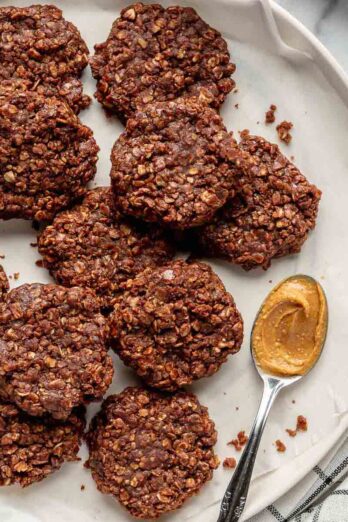 Batch of no-bake oatmeal cookies on a parchment paper-lined plate with a spoon of peanut butter.