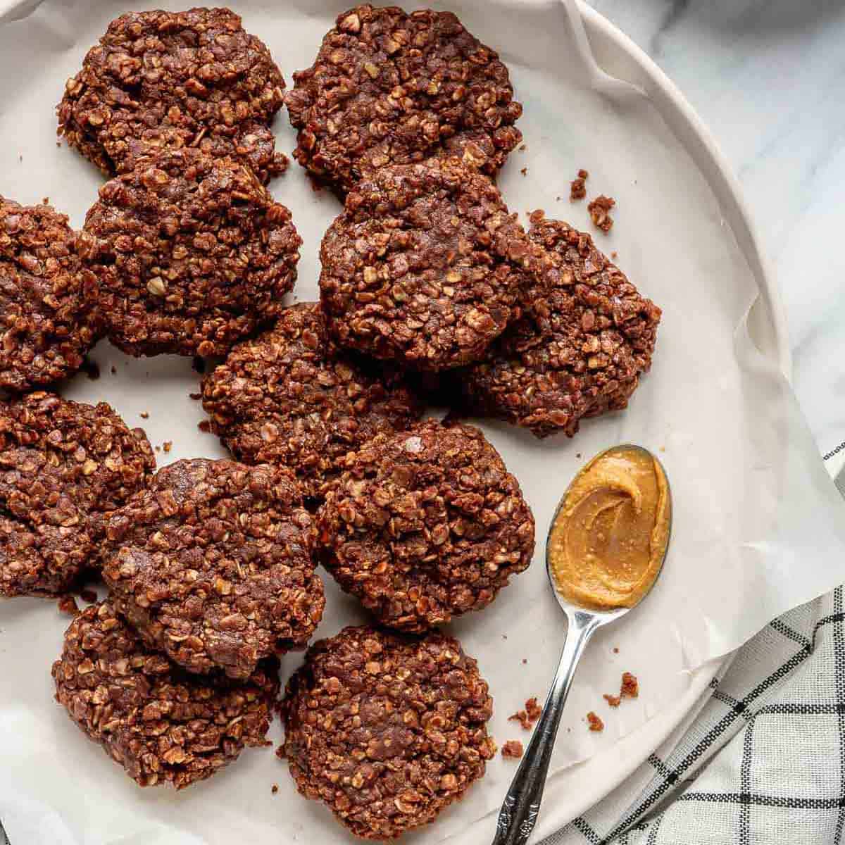 Batch of no-bake oatmeal cookies on a parchment paper-lined plate with a spoon of peanut butter.