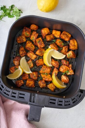 Air fried salmon bites in an air fryer basket garnished with fresh chopped herbs and lemon wedges.