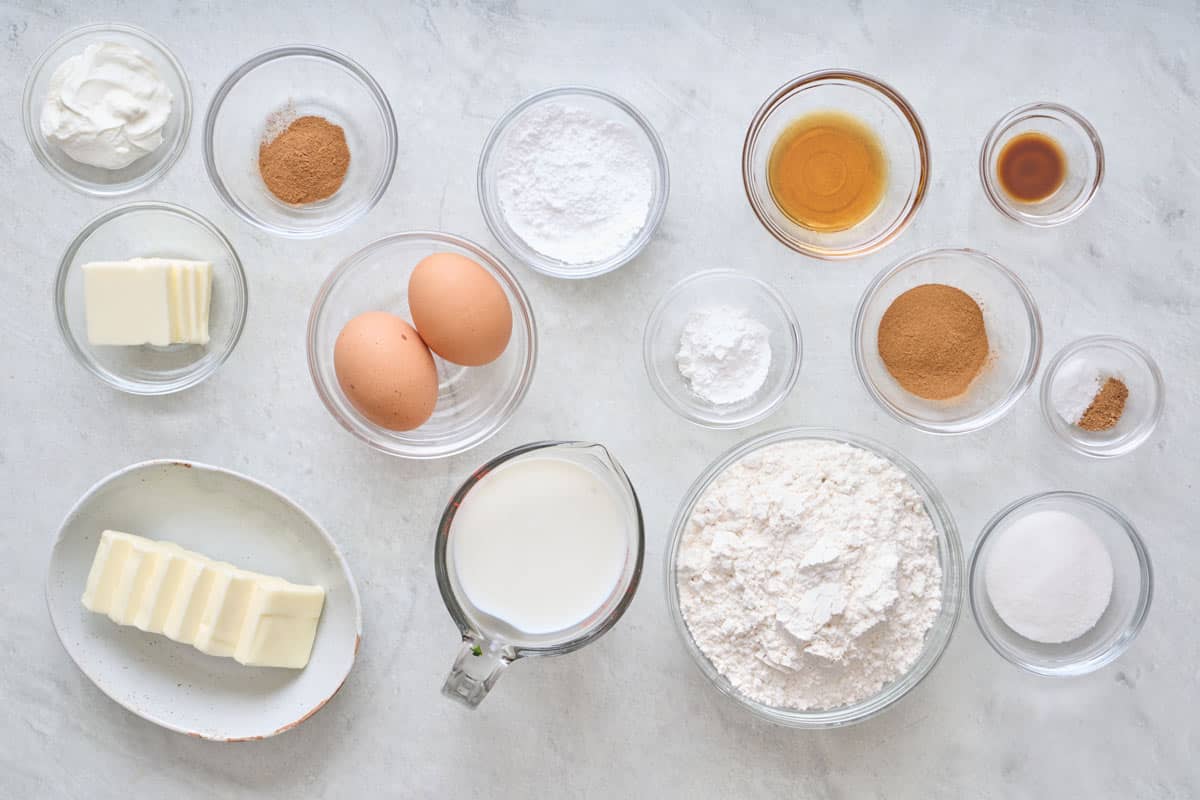 Ingredients for recipe in individual bowls: Greek yogurt, butter, cinnamon, eggs, nut meg, sugar, milk, vanilla powdered sugar, baking powder, and flour.