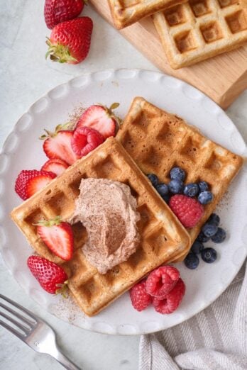 Two cinnamon waffles on a serving plate, topped with whipped cinnamon butter and fresh fruit.
