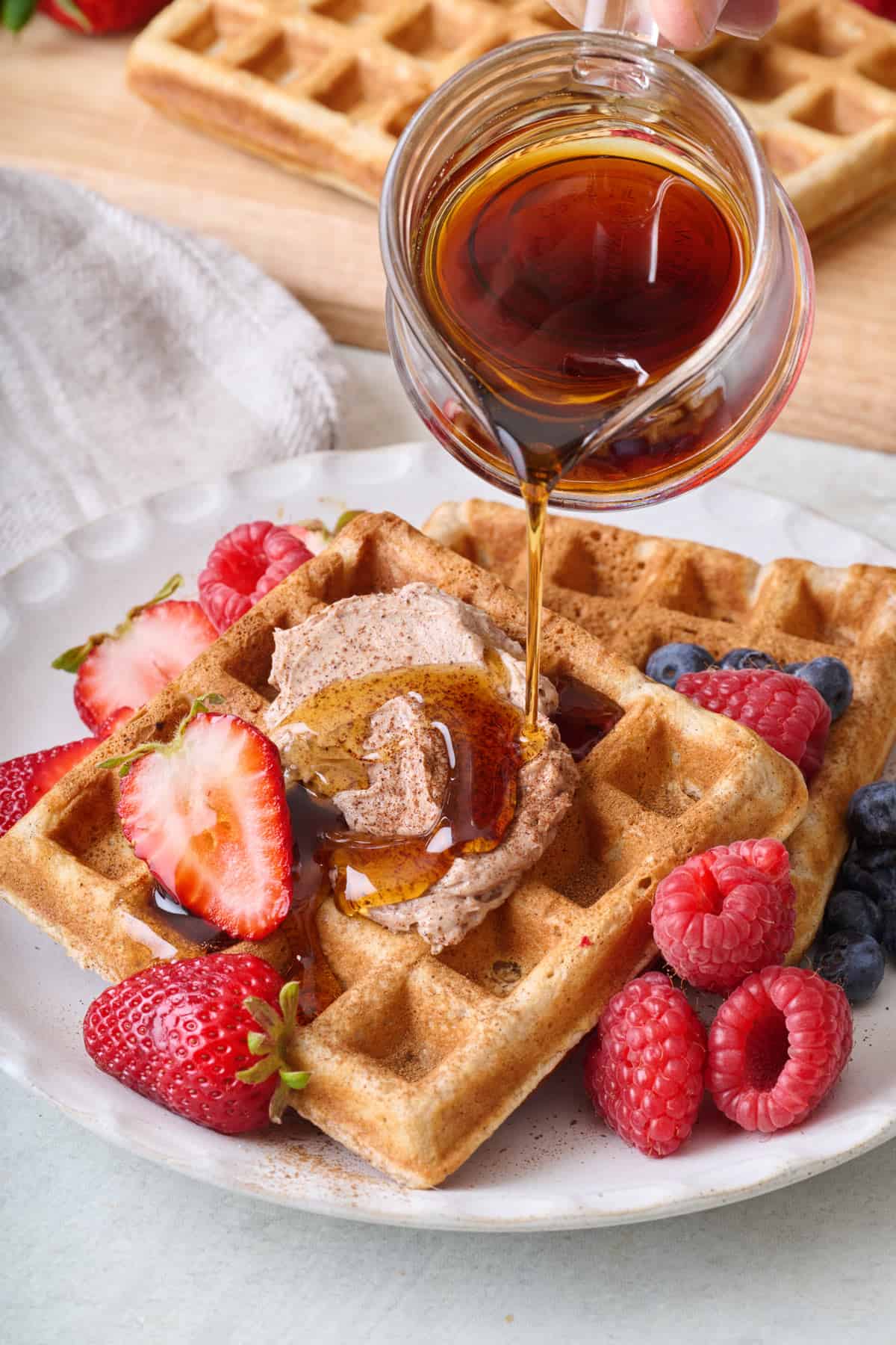 Maple syrup being poured over a serving of cinnamon waffles with whipped butter and fresh fruit.