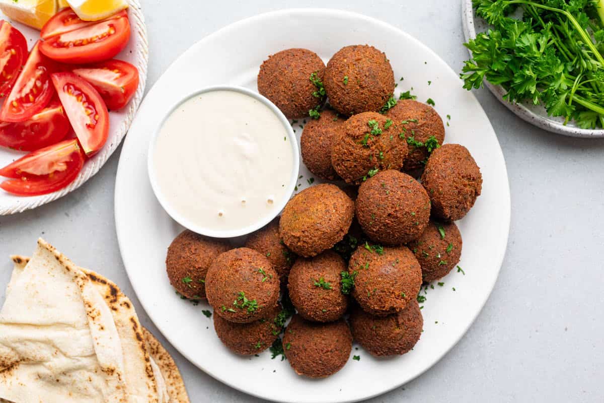 Falafel on a plate with a tahini dip surrounded by pita bread, tomatoes and greens.