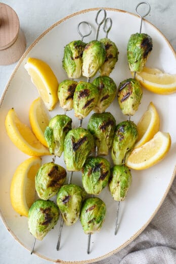 Grilled brussel sprouts on metal skewers, served on a large platter with lemon wedges.