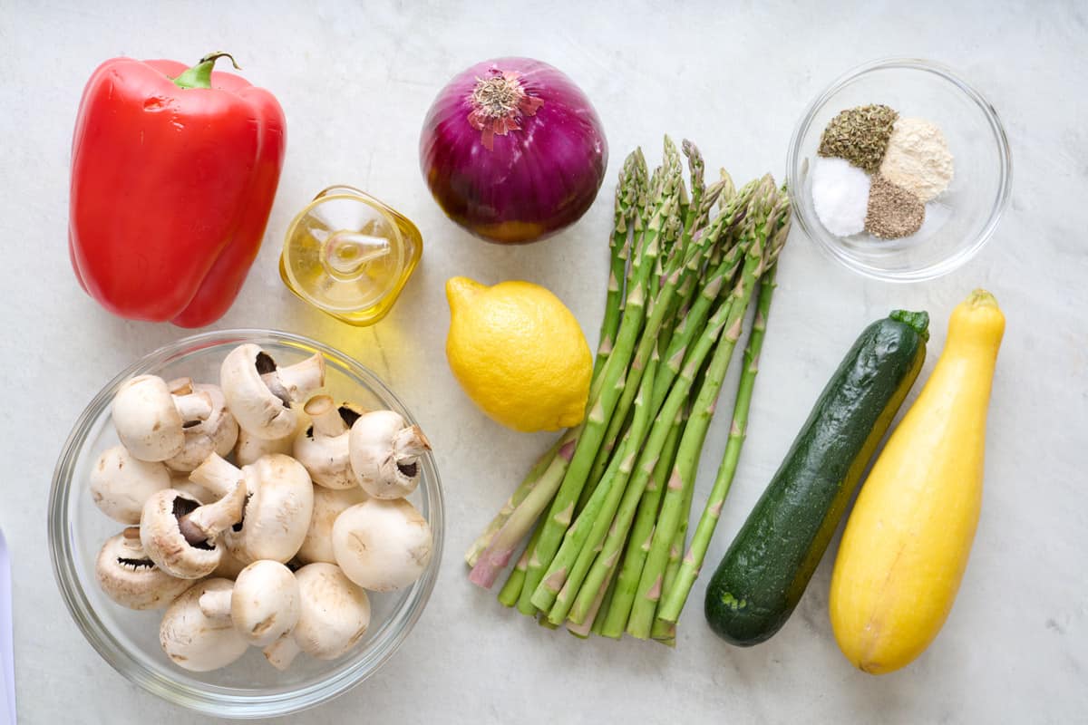 Ingredients for recipe before prepping: red bell pepper, mushrooms, oil, red onion, lemon, asparagus, zucchini, squash, and seasoning.