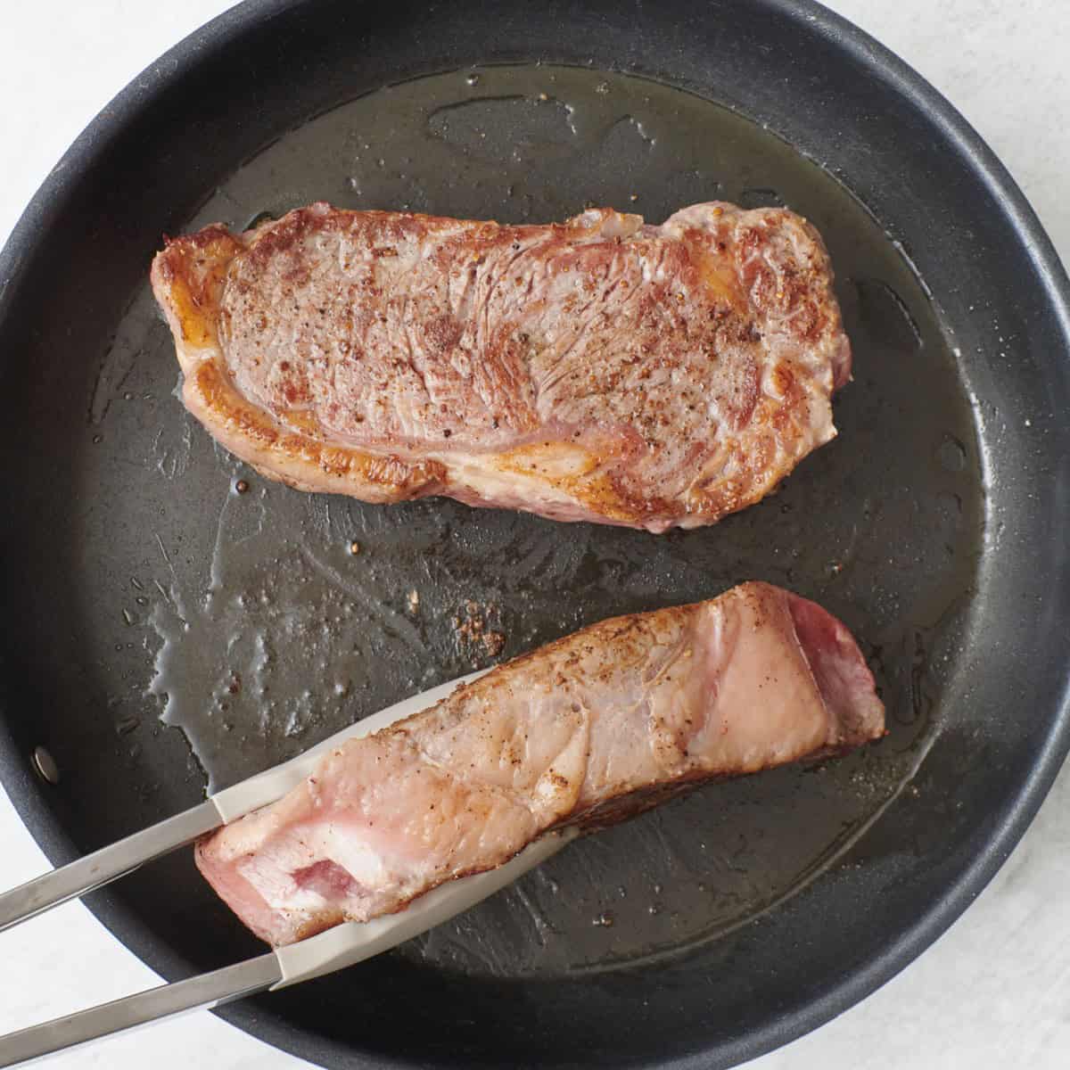 One steak flat down and the other one on its side held up with tongs.