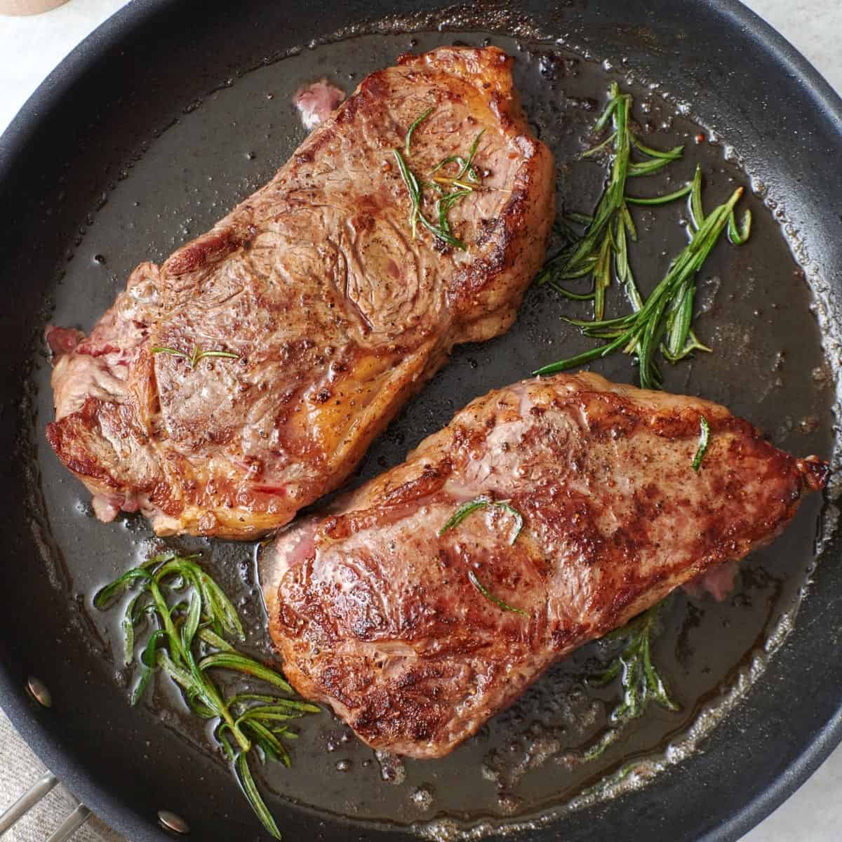 NY Strip Steak on a non-stick skillet with melted butter spooned on top and fresh thyme leaves on top.