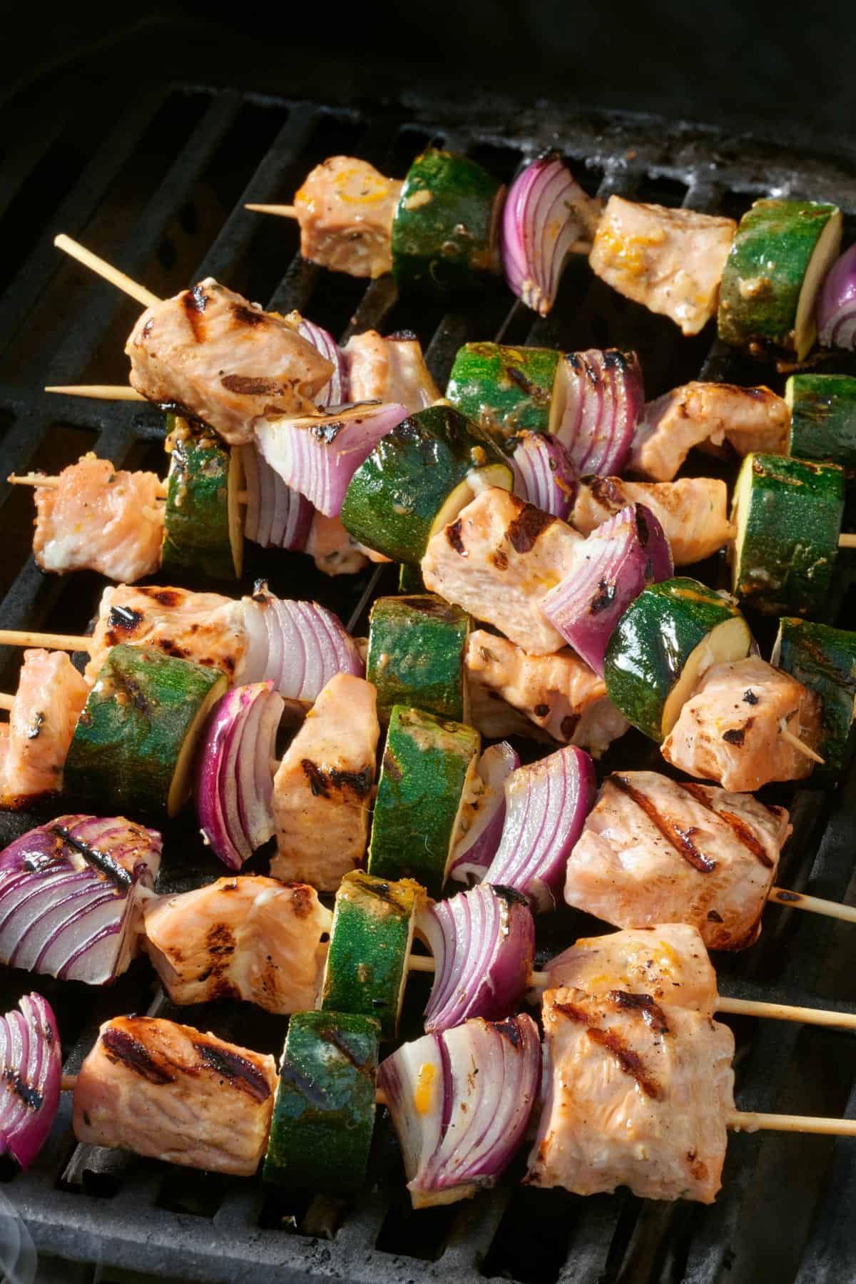 Pile of salmon kabobs skewered with red onions and zucchini on the grill with grill marks.