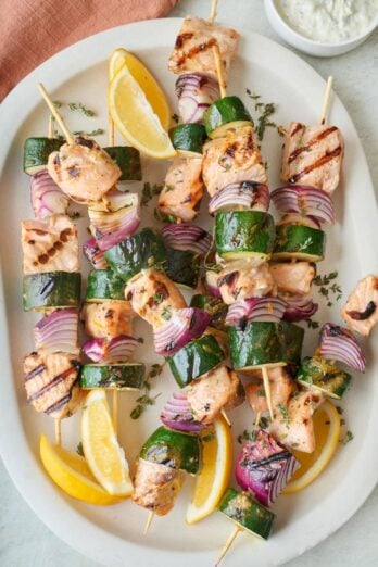 Salmon kabobs with vegetables on a large platter, garnished with fresh herbs and lemon wedges.