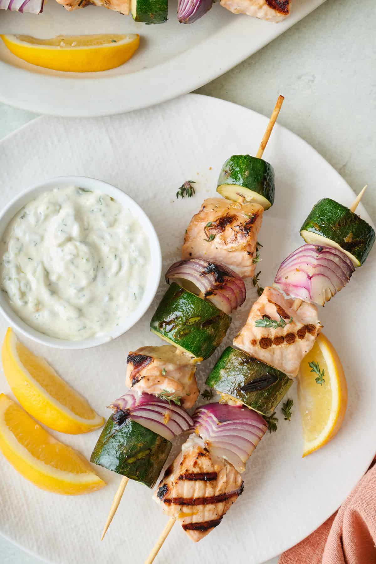 Two salmon skewers on a plate with homemade tartar sauce and lemon wedges.