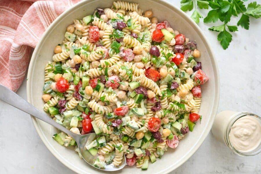 Tahini pasta salad in a large bowl with dressing and ingredients nearby.