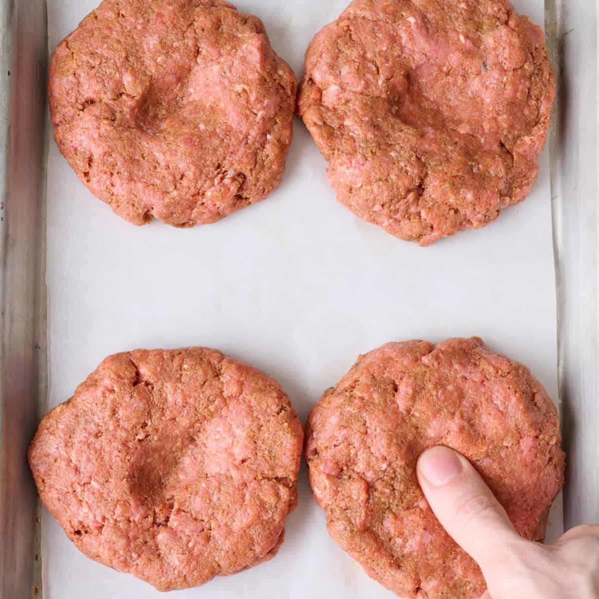 A person using their thumb to make indents in the centers of four hamburger patties.