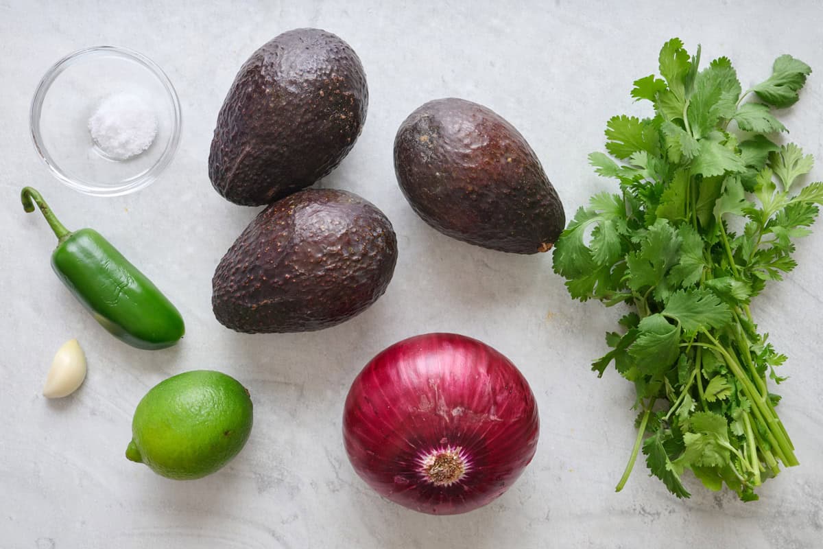 Avocados, lime, cilantro, red onion, jalapeno, garlic, and salt on a marble countertop.