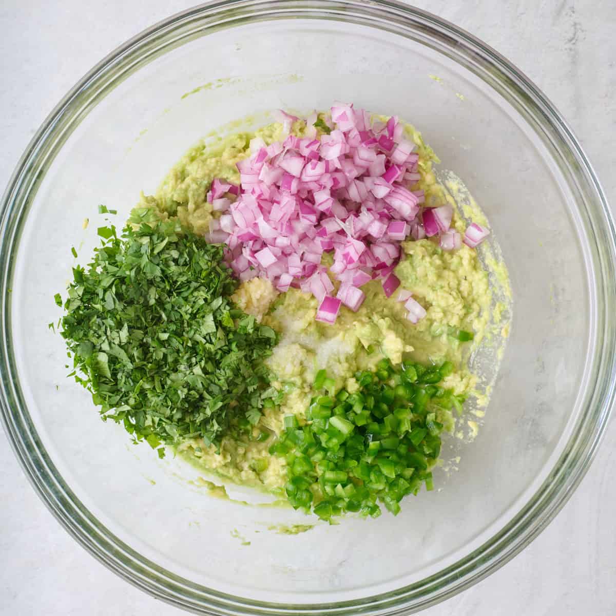 Mashed avocado and minced jalapeno, red onion, and cilantro in a large glass mixing bowl.