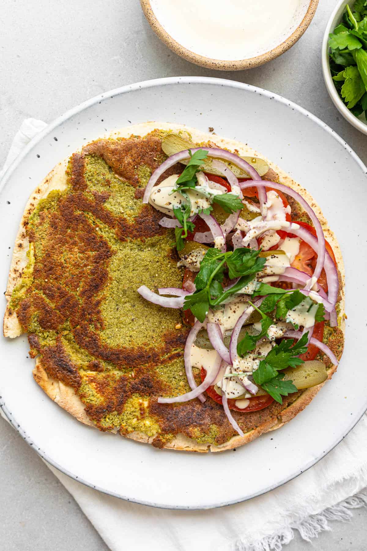 Smash falafel pita open-faced sandwich on a plate with tomatoes, red onions, parsley, pickles and tahini sauce drizzled on top