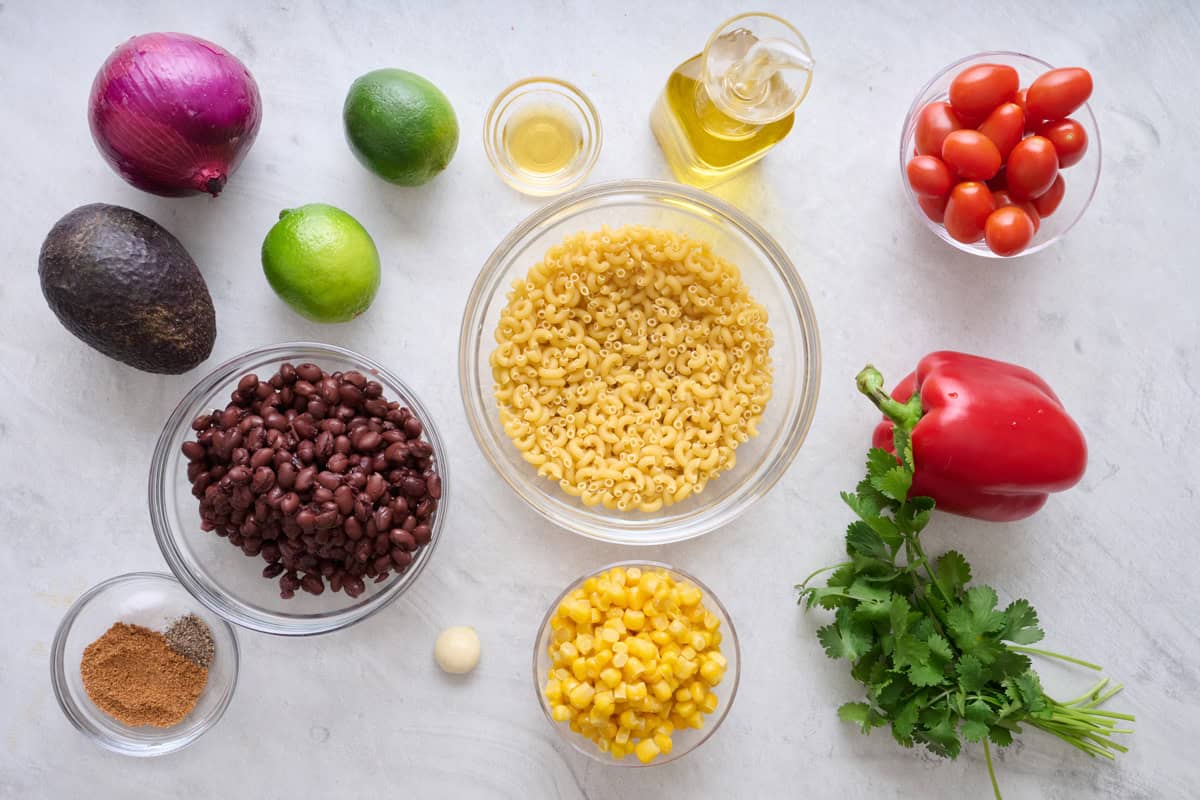 Taco pasta salad ingredients on a marble countertop: noodles, black beans, veggies, cilantro, limes, and spices.