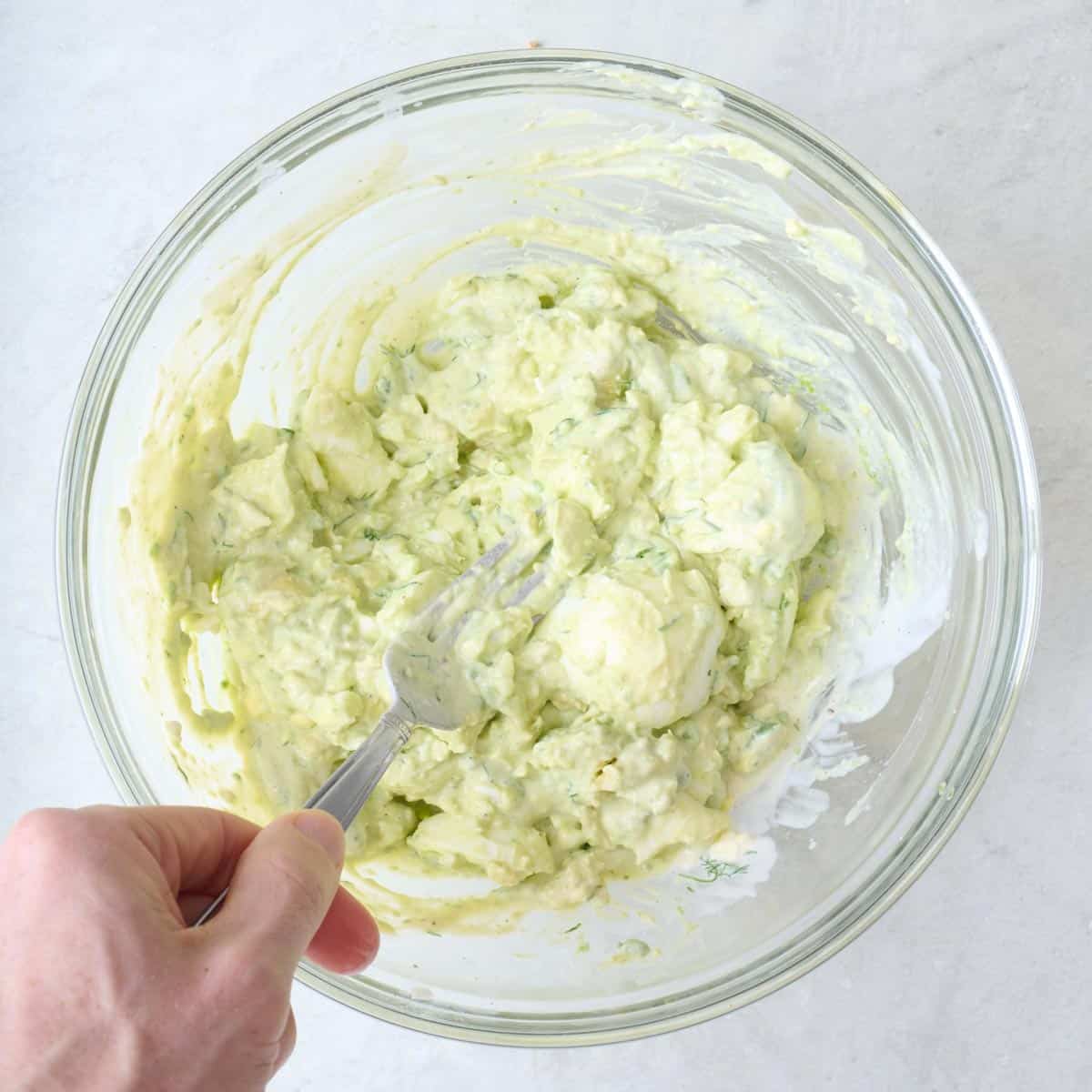 Fork mashing boiled eggs, avocado, and dressing together.