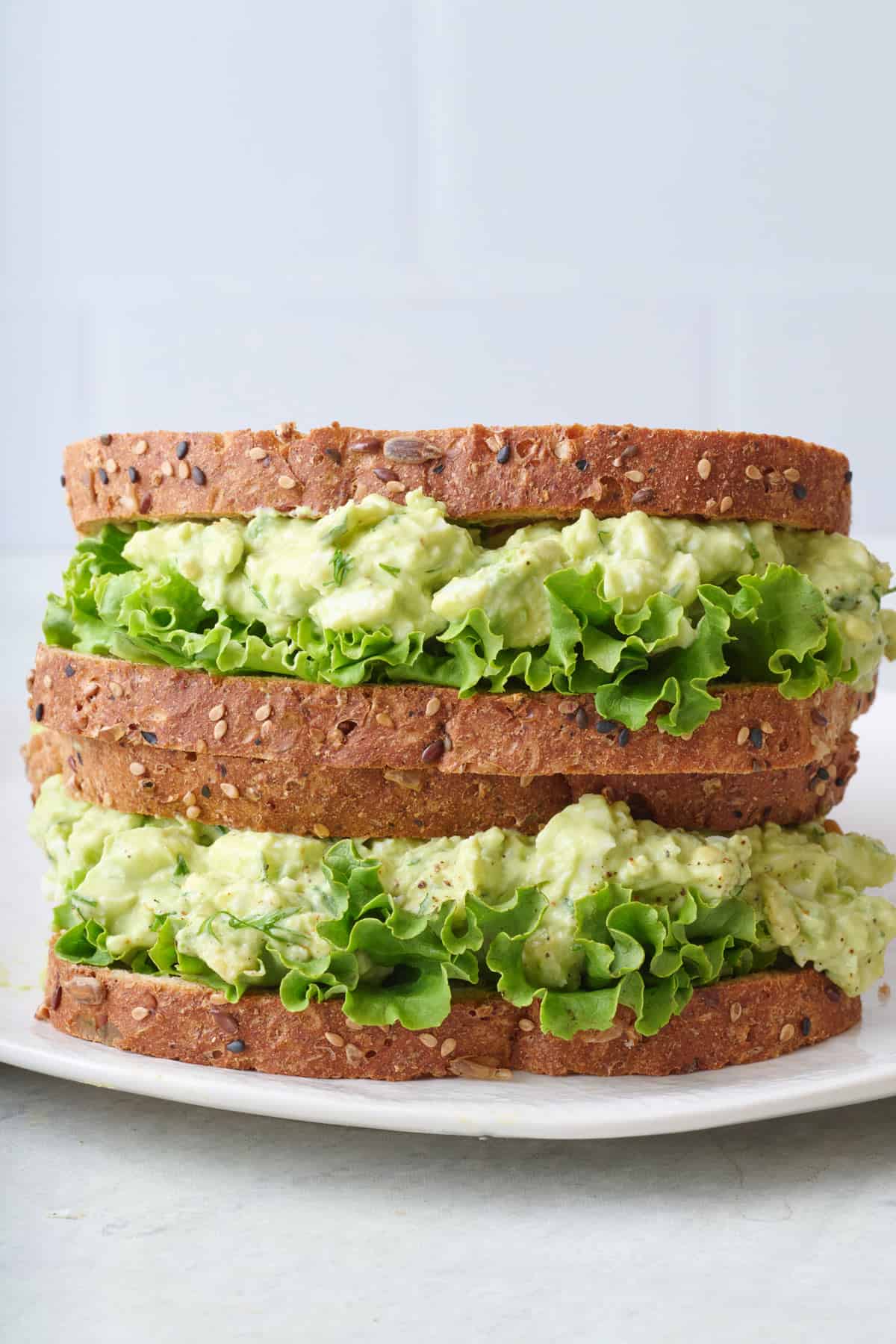 Two avocado egg salad sandwiches with lettuce stacked on top of each other on a plate.