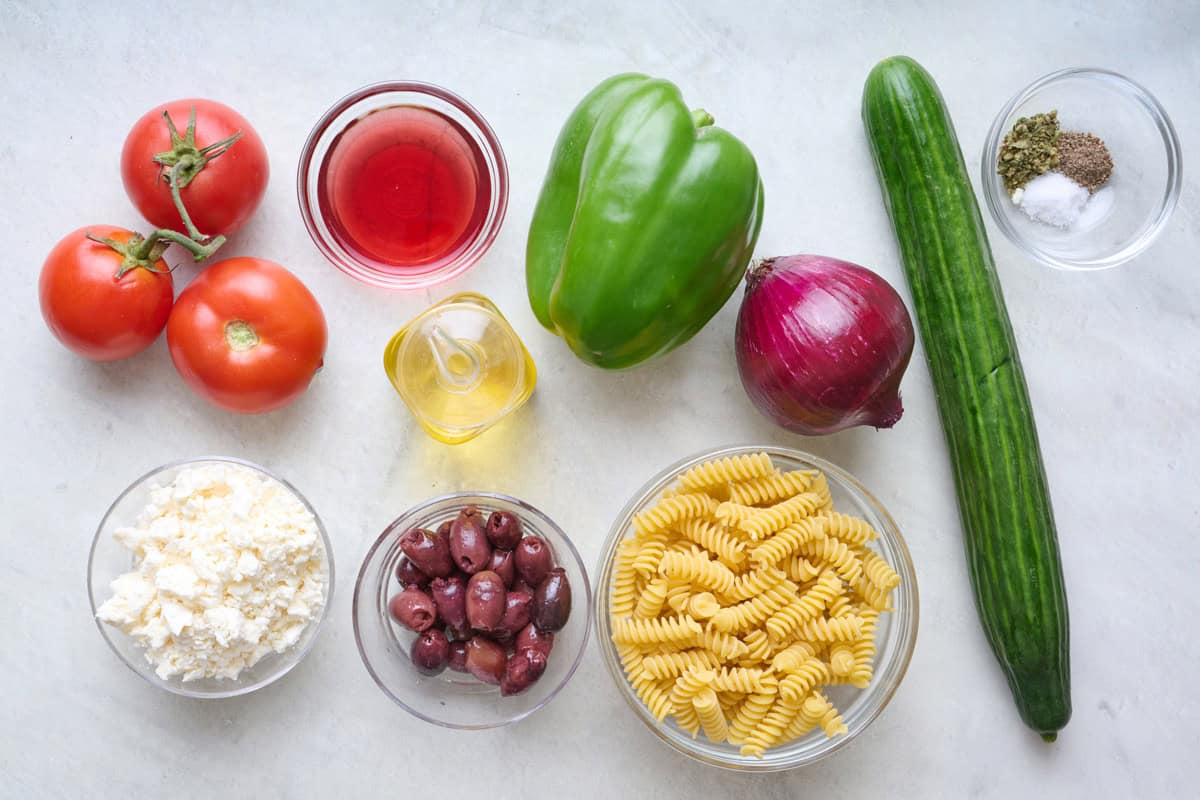Ingredients for Greek pasta salad, including tomatoes, cucmber, dried pasta, olives and feta cheese.