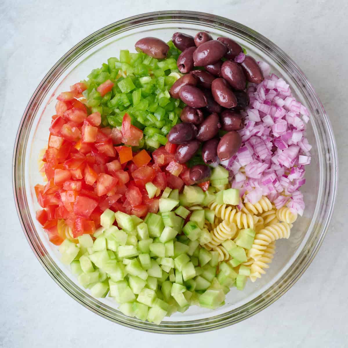 Chopped cucumber, tomatoes, bell pepper and red onion, olives, and cooked pasta in a large glass mixing bowl.