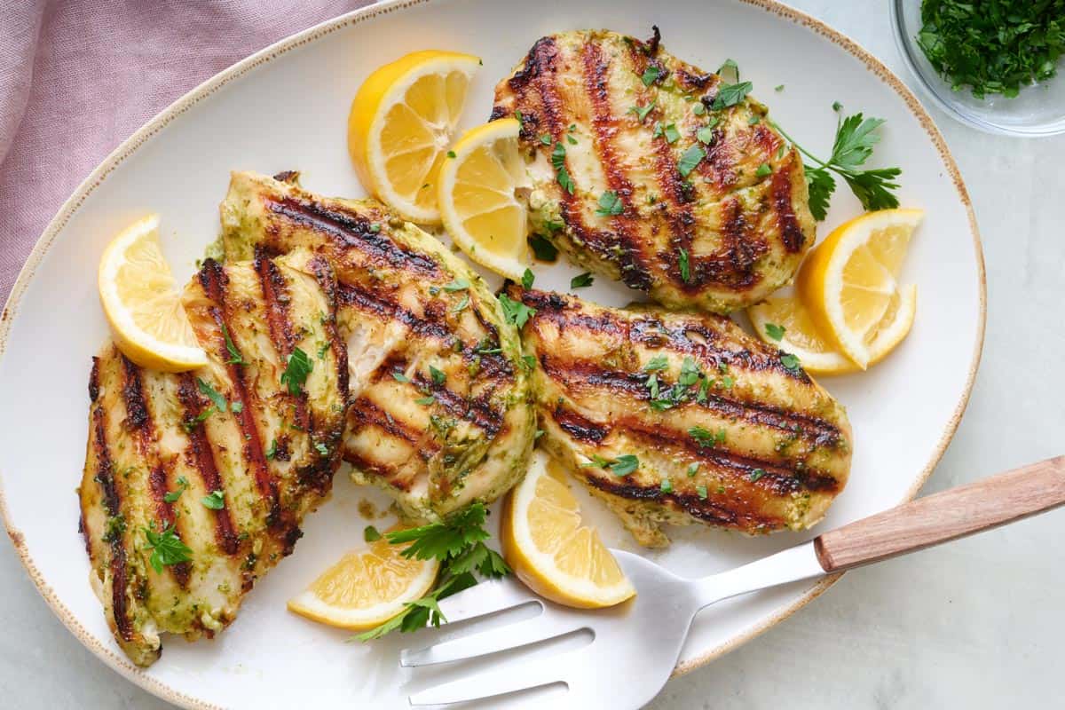 Pesto grilled chicken breasts on a platter, garnished with lemon wedges and fresh parsley.
