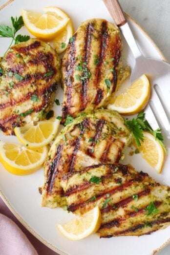 Pesto grilled chicken breasts on a platter, garnished with lemon wedges and fresh parsley.