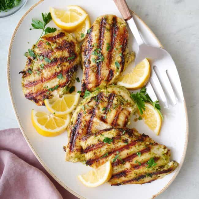 Pesto grilled chicken breasts on a platter, garnished with lemon wedges and fresh parsley.