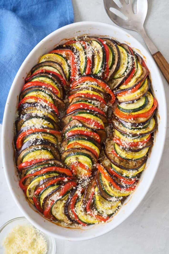 Ratatouille in baking dish garnished with red wine vinegar and freshly shredded parmesan cheese with serving spoon nearby.