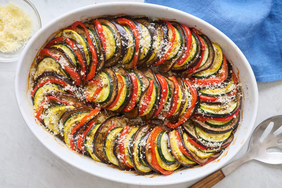 Ratatouille in baking dish garnished with red wine vinegar and freshly shredded parmesan cheese with serving spoon nearby.