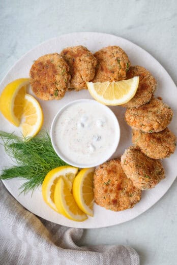 Salmon patties around the edge of a round plate with a bowl of yogurt sauce in the middle for dipping, garnished with fresh dill and lemon wedges.