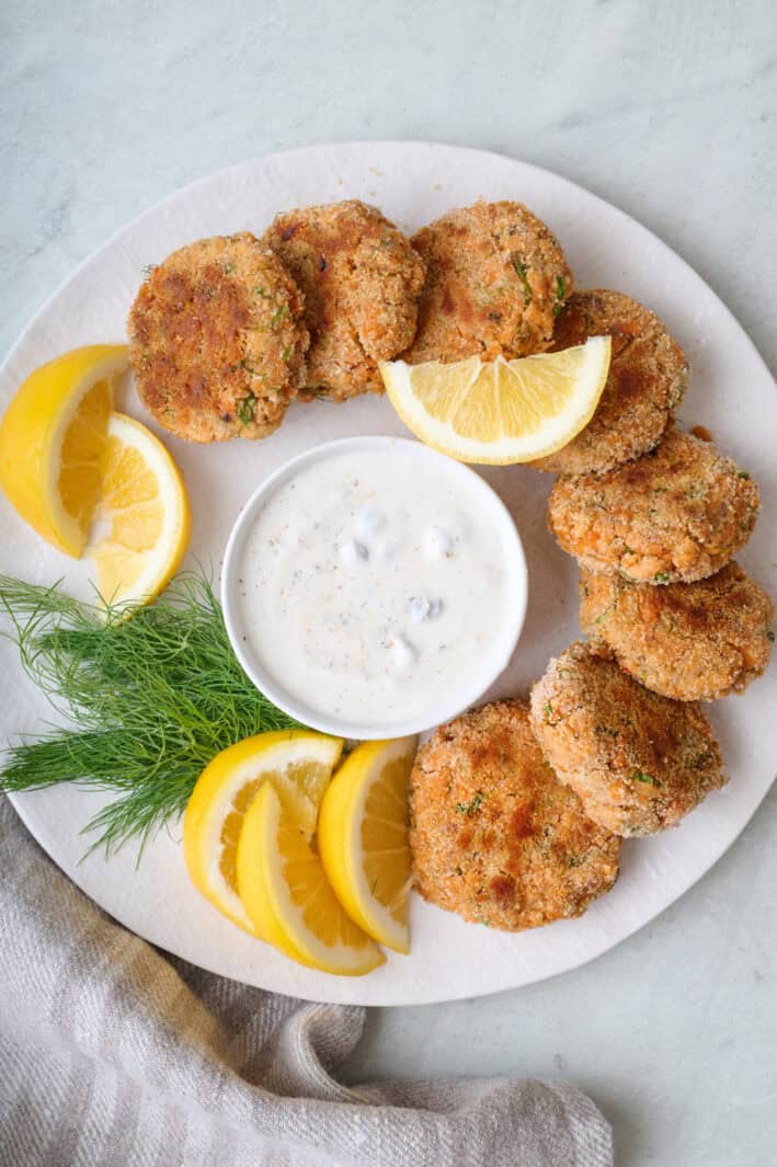 Salmon patties around the edge of a round plate with a bowl of yogurt sauce in the middle for dipping, garnished with fresh dill and lemon wedges.