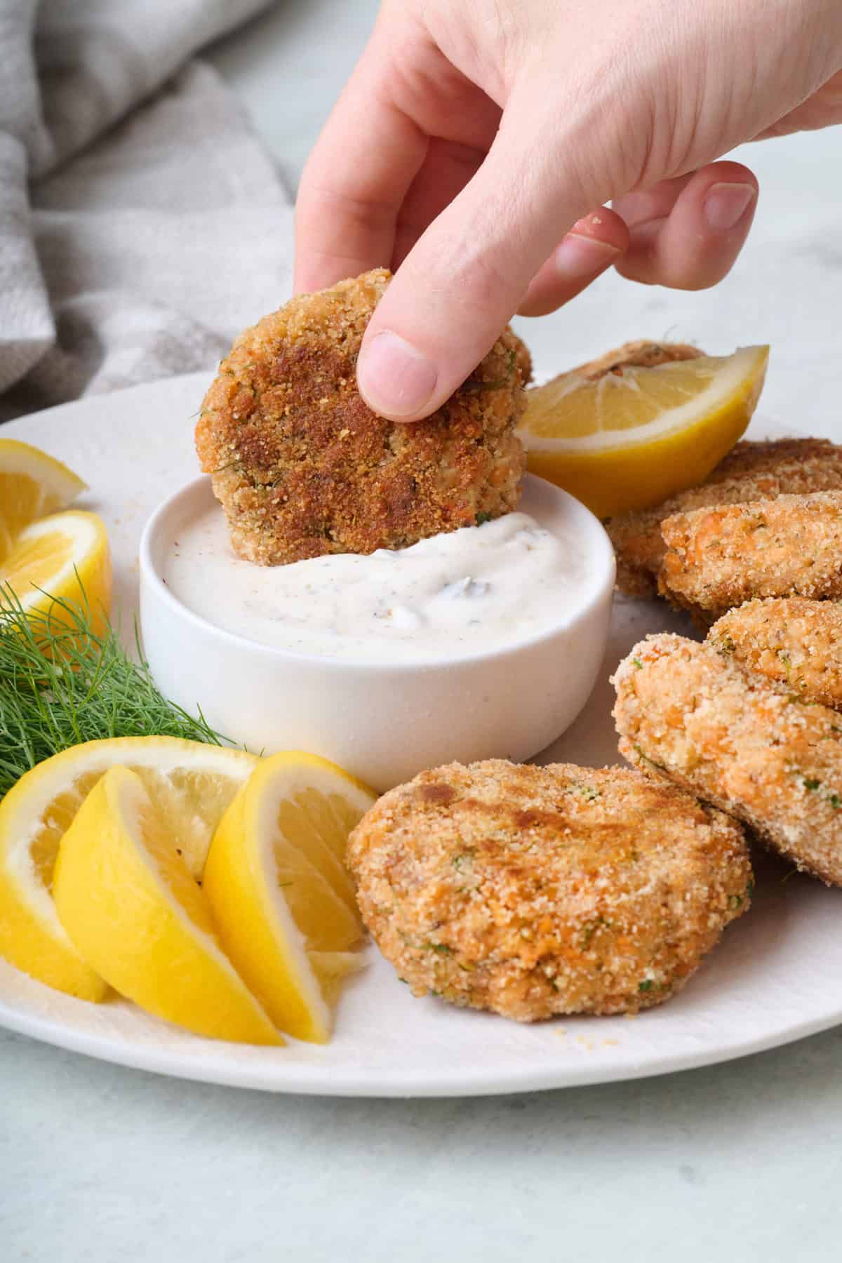 Hand dipping a salmon patty into Greek yogurt sauce with more patties on platter.