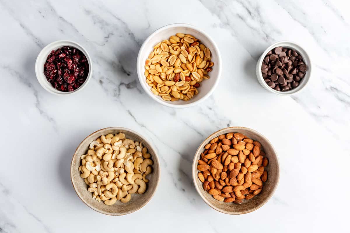 Small bowls of dried cranberries, peanuts, chocolate chips, cashews, and almonds on a marble countertop.