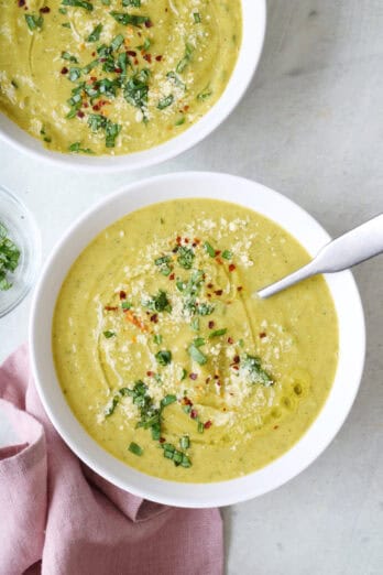 Two bowls of blended zucchini soup garnished with oil, parmesan, red pepper flakes and basil.