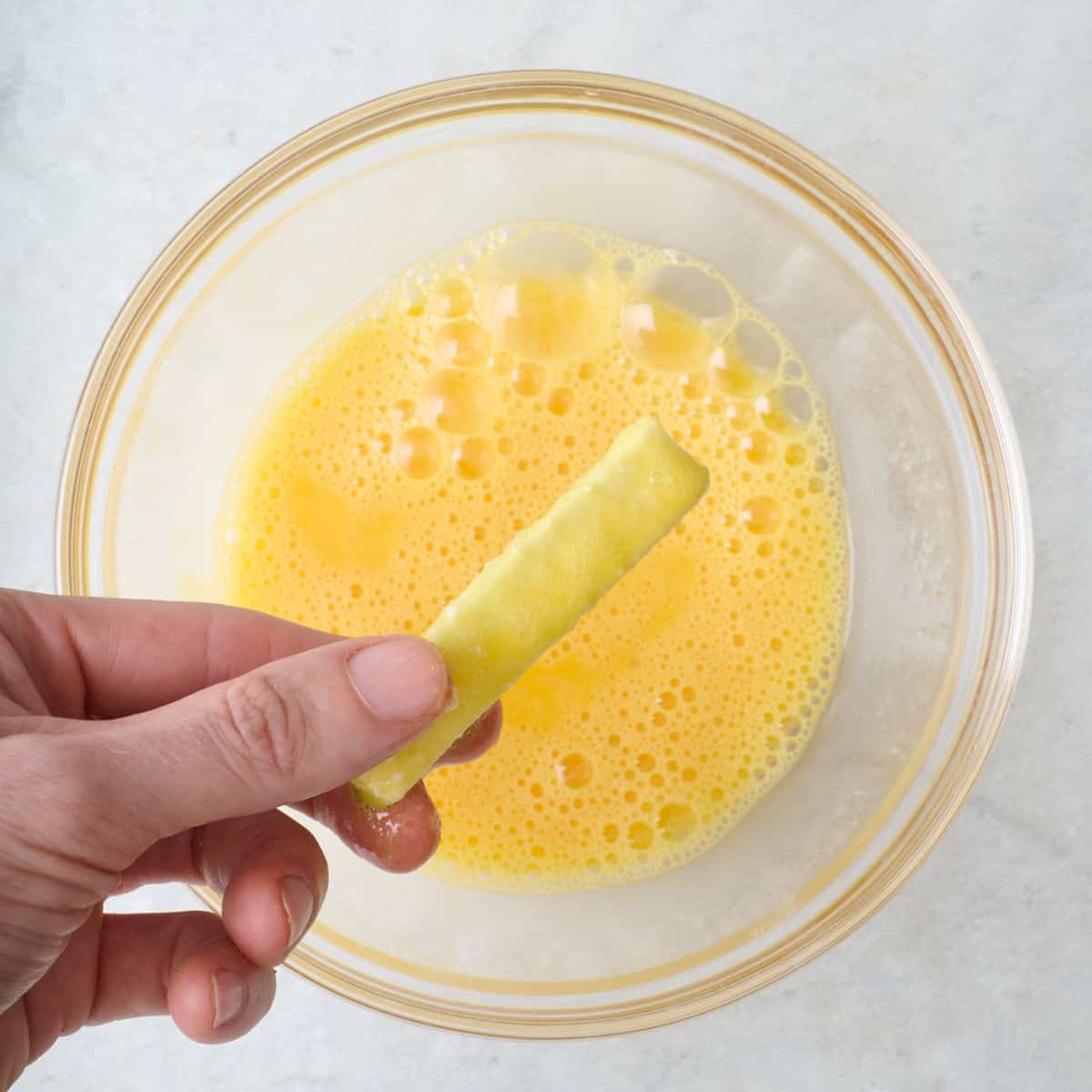 A zucchini stick being dipped into beaten eggs in a bowl.