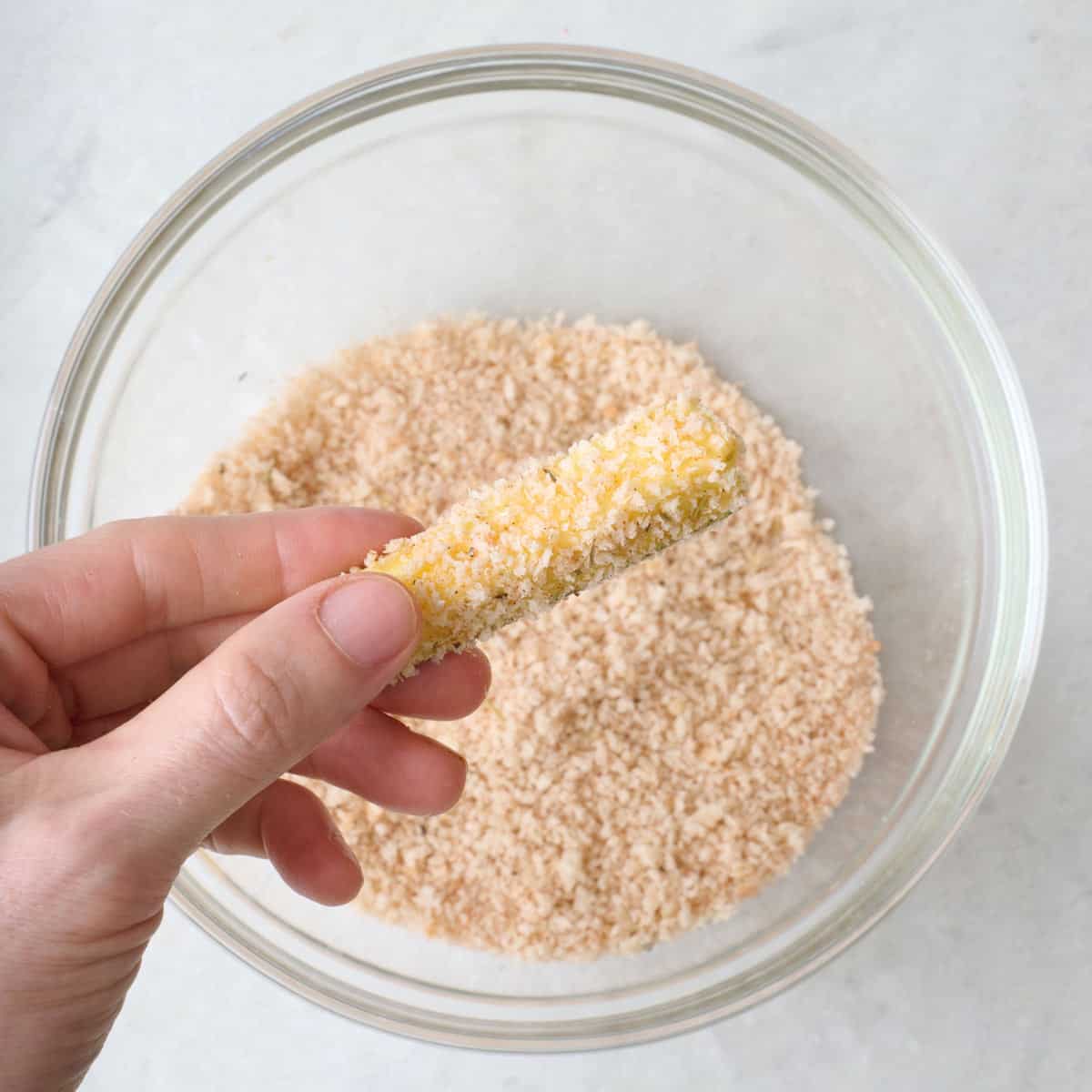 A zucchini stick being dipped into a breadcrumb mixture in a bowl.